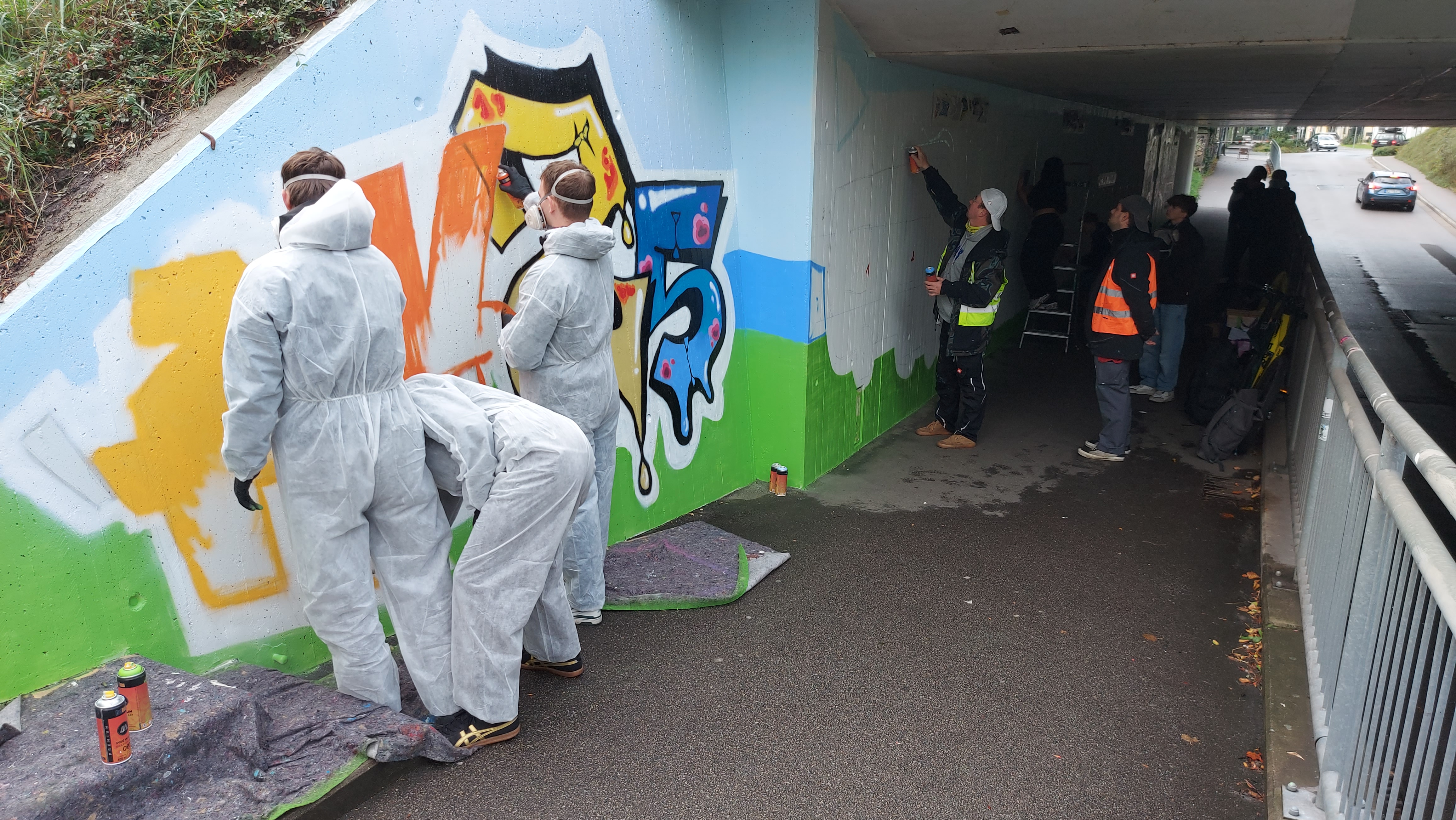 Graffitisprüher bei der Arbeit. Schüler in weißen Schutzoveralls vor ihrem Bild.