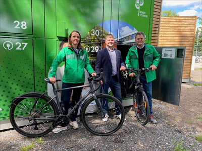 Drei Personen stehen mit Fahrrädern vor grünen Fahrradboxen.